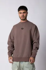 Buzo de cuello redondo en color marrón, con diseño oversize y mangas largas. Presenta un pequeño logo circular con las siglas VCP en el centro del pecho y un detalle de etiqueta pequeña en el puño.