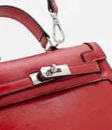 Cartera pequeña de poliuretano color rojo, con diseño estructurado, asa superior rígida y cierre frontal con herraje metálico.