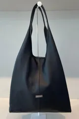 Bolso tipo shopper de gran capacidad, confeccionado en cuero vacuno negro. Presenta un diseño minimalista con costura central vertical y placa metálica con el logo de la marca. Cuenta con fuelles laterales ajustables mediante cierres para ampliar su volumen, asas integradas y un sobre de cuero desmontable en su interior.