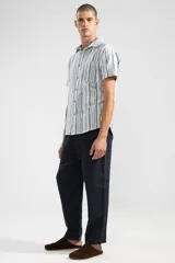 Camisa de manga corta con rayas verticales en tonos azul, gris y blanco. Tiene cuello clásico, botones de coco y un bolsillo en el pecho.