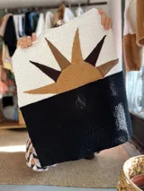 Buzo de tejido tipo corderito con diseño de atardecer en el frente, combinando bloques de color blanco y negro con apliques en tonos tierra. Presenta cuello redondo y mangas largas.