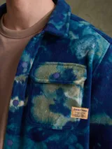 Sobrecamisa de tejido polar con estampado abstracto en tonos rojizos y azul marino. Presenta cuello clásico, cierre frontal con botones a presión, dos bolsillos frontales con solapa en el pecho y un parche decorativo.