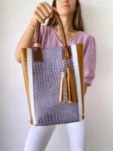 Bolso tipo tote de cuero color lila con textura croco, detalles en cuero marrón y laterales con franjas blancas. Incluye manijas de cuero marrón y accesorio de borlas colgantes.