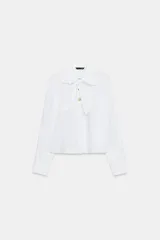Camisa blanca de cuello solapa y manga larga, con detalle de lazada en el mismo tejido en la parte frontal y cierre con botones dorados.