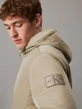 Campera Calvin Klein color verde oliva, con capucha, cierre frontal con cremallera, puños y bajo elásticos, bolsillos con botón de presión en la cintura y logo de la marca en el pecho y la manga. Confeccionada en nailon 100% reciclado y forro de poliéster.