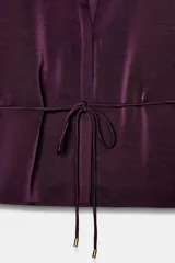 Camisa satinada de color violeta oscuro, con cuello solapa, escote en pico y manga larga. Presenta un detalle de cinturón fino ajustable a tono en la cintura y cierre frontal con botones.