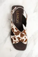 Sandalias bajas con tiras cruzadas de cuero con estampado de vaca en tonos marrón y blanco. Tira trasera con hebilla dorada y suela plana color marrón.