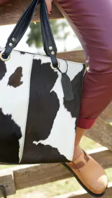 Cartera tipo tote bag de cuero nonato con estampado de vaca en blanco y negro, doble asa corta de cuero negro y etiqueta colgante con el nombre de la marca.