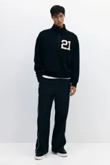 Buzo negro de tejido pesado con interior cepillado suave, cuello elevado con medio cierre y hombros ligeramente caídos. Presenta un estampado frontal con el número "21" en color beige. Puños y bajo en punto acanalado.