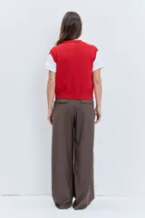 Chaleco de punto rojo con cierre frontal de botones, cuello redondo y dos bolsillos frontales con solapa. Presenta un diseño de tejido acanalado en los bordes y un pequeño parche decorativo en el pecho.