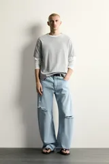 Jeans de corte barrel fit confeccionados en denim de algodón rígido, con diseño de cinco bolsillos y cierre frontal con botones. Presentan un lavado claro con efecto de roturas en las perneras.