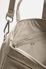 Bolso tipo shopper de tejido técnico color beige, con dos asas de mano y una correa de hombro ajustable y extraíble. Presenta un compartimento principal con cierre de cremallera, un bolsillo interior con cremallera y dos bolsillos frontales con cierre de botón a presión.