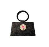 Bolso de mano tipo sobre, confeccionado en tejido negro con textura, con asa circular rígida recubierta de mostacillas negras. Presenta un aplique circular metálico dorado con una piedra de resina rosada en el centro.