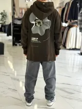 Canguro oversize chocolate con capucha, confeccionado con un diseño urbano que incluye un gran estampado gráfico en la espalda con líneas onduladas y texto.
