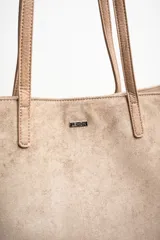 Bolso tipo tote de gran tamaño confeccionado en gamuza sintética color tostado. Presenta un diseño minimalista con asas largas de hombro y una pequeña placa metálica frontal con el logo de la marca.
