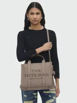 Cartera tipo tote de cuero granulado color tostado, con diseño estructurado y rectangular. Presenta doble asa superior fija y una correa larga ajustable y extraíble. En el frente, destaca el logo estampado con la inscripción 'THE TOTE BAG MARC JACOBS'.