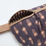 Riñonera acolchada de color púrpura con estampado floral en tonos claros. Presenta cierre metálico dorado, correa ajustable de tela en color beige y herrajes metálicos.