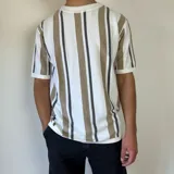 Remera de tejido de punto en hilo de algodón, con diseño de rayas verticales en tonos blanco, beige y gris oscuro. Presenta cuello redondo y mangas cortas con terminaciones acanaladas, de corte semi oversize.