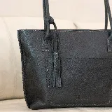 Cartera tipo tote de cuero marrón con costuras artesanales visibles, bolsillo frontal con el logo grabado, asas de hombro y detalles de flecos colgantes.