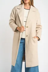 Tapado largo de corte oversize en color beige, con solapas clásicas y cierre frontal con botones. Presenta un diseño minimalista y elegante, ideal para un look sofisticado.
