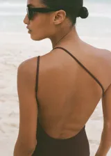 Traje de baño enterizo marrón chocolate con diseño drapeado, cuello asimétrico con tirante fino y aro decorativo dorado.