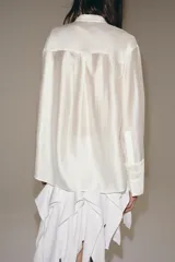 Camisa de cuello mao color beige, confeccionada en tejido satinado con brillo sutil, manga larga con puños anchos y un bolsillo de parche en el pecho con detalles bordados tonales. Cierre frontal con botones ocultos bajo solapa.