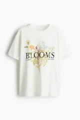 Remera de algodón color blanco crudo, corte oversized y cuello redondo. Presenta un estampado central de flores silvestres con la palabra "BLOOMS" en tipografía serif negra, y las palabras "FLORIST" y "HARVESTS" a los lados.