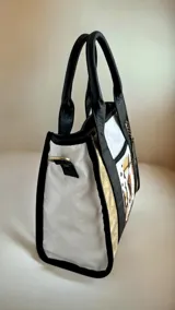 Cartera de cuero modelo Tali, de diseño estructurado que combina los colores blanco, negro y dorado, con detalles de arte pintado a mano en el frente. Cuenta con doble asa de mano, correa larga desmontable y herrajes metálicos.
