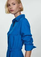 Vestido midi de corte camisero color azul vibrante, confeccionado en mezcla de lyocell y lino. Presenta cuello camisero, cierre frontal con botones, mangas largas con puños abotonados y dos bolsillos de parche en el pecho. Incluye un lazo ajustable en la cintura para entallar la silueta y aberturas laterales en el bajo.
