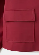Chaqueta corta de algodón en color rojo, con cuello camisero, manga larga y cierre frontal de botones oscuros. Presenta dos bolsillos con solapa en el frente.