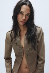 Blazer corto de estilo militar color tostado, con cuello redondo, manga larga y cierre frontal con ganchos metálicos. Presenta detalle de botones decorativos dorados en el frente y en los hombros, con costuras marcadas y forro interior a contraste.