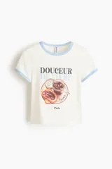 Remera corta de algodón blanca con ribetes celestes en el cuello y las mangas. Presenta un estampado frontal con la inscripción "Douceur de vivre" y una ilustración de tostadas con mermelada y un frasco.