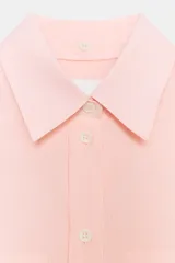 Camisa de corte oversize color rosa claro con diseño de rayas verticales sutiles. Presenta cuello solapa desmontable, cierre frontal con botones ocultos bajo solapa, mangas largas con puños y bajo asimétrico redondeado. Incluye dos bolsillos frontales amplios.