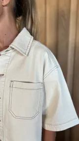 Sobrecamisa de denim soft en tono azul medio, con corte oversize, mangas cortas, cuello clásico y cierre frontal con botones metálicos. Presenta dos bolsillos frontales con solapa y un ruedo redondeado.