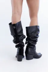 Bota de cuero negro de caña alta slouchy, con diseño fruncido ajustable mediante un juego de correas y hebillas. Presenta taco bajo cuadrado y punta cuadrada.