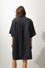 Vestido camisero corto en denim color gris oscuro, de corte holgado, con cuello camisero, manga corta, bolsillo frontal y cierre frontal con botones blancos.