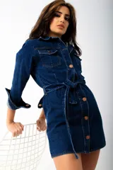 Vestido camisero corto de denim azul oscuro, con cuello clásico, mangas 3/4 con puños abotonados, cierre frontal con botones color cobre, cinturón de lazo y bolsillos con solapa en el pecho.