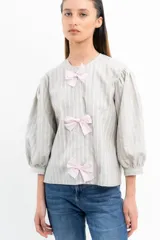 Blusa a rayas verticales grises y blancas con mangas abullonadas y tres lazos rosas en el frente.