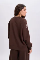 Sudadera de cuello redondo y manga larga en color marrón, con diseño holgado y detalle de etiqueta en la manga.
