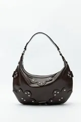 Cartera tipo hobo de símil cuero texturizado color marrón oscuro, con apliques de tachas metálicas plateadas y detalles de recortes florales en el frente y los laterales. Cuenta con asa corta para llevar al hombro y cierre superior.