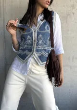 Chaleco de jean azul con bordados florales blancos y ribete de encaje. Cierre frontal con botones.