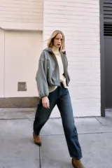 Chaqueta estilo bomber de corte oversize en color marrón oscuro, confeccionada en tejido suave. Presenta capucha con cordones ajustables, cierre frontal con cremallera, mangas largas con puños elásticos y bolsillos delanteros de vivo. El bajo cuenta con detalle de pliegues.