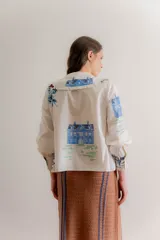 Camisa de popelina de algodón color crudo, con cuello bobo amplio con volados y cierre frontal de botones. Presenta un diseño estampado con motivos botánicos y de aves en tonos azules y verdes.