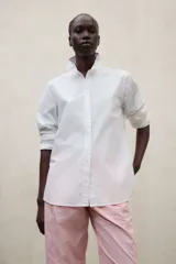 Camisa blanca de corte holgado, con cuello camisero rígido, cierre frontal de botones ocultos y manga larga.