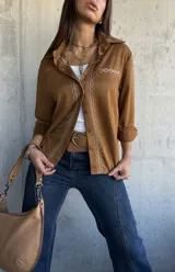Camisa de gamuza color camel, con cuello clásico, cierre frontal de botones y detalle de bordado geométrico en color crema a lo largo de la botonadura y en el bolsillo frontal. Presenta mangas largas con puños doblados.