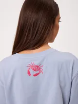 Remera celeste de corte holgado, con cuello redondo, manga corta y estampado frontal colorido de temática costera.