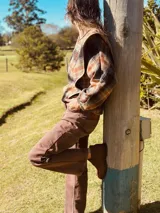 Camisa manga larga con estampado a cuadros en tonos marrón, naranja y negro, con cuello clásico y cierre frontal con botones. Presenta volados en delantero y espalda.
