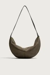 Bolso de hombro tipo hobo con diseño de media luna, confeccionado en gamuza color marrón. Cuenta con un asa larga fija de cuero negro y cierre superior con cremallera.