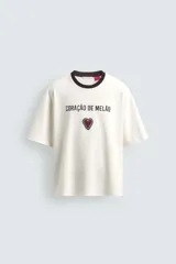 Camiseta de corte relajado confeccionada en algodón, color blanco crudo con cuello redondo en contraste color negro. Presenta un bordado frontal con la frase 'Coração de Melão' y un parche en forma de corazón.