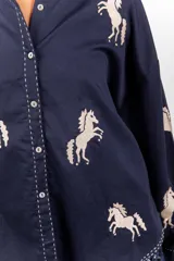 Camisa de corte clásico y calce amplio, color marfil, con cuello camisero, mangas largas y cierre frontal con botones. Presenta bordados coloridos de caballos en contraste distribuidos en la prenda.